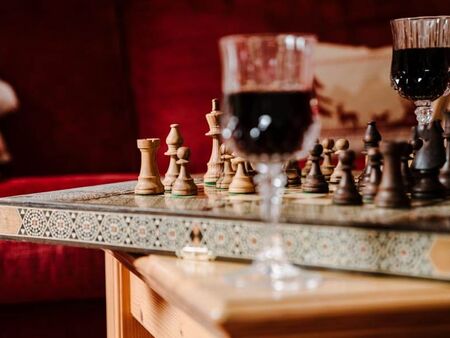 Ein kunstvoll verziertes Schachbrett steht auf einem hölzernen Tisch, daneben stehen zwei Weingläser mit Rotwein und im Hintergrund ist eine rote Samtcouch
