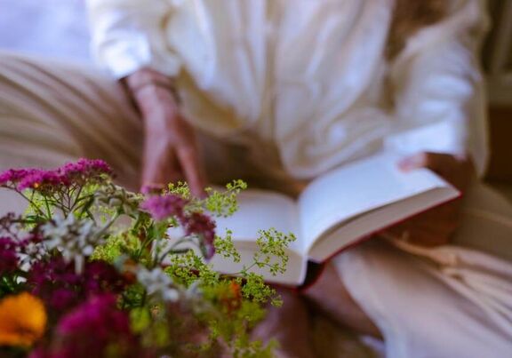 An elderly lady reading a book next to a flower bush