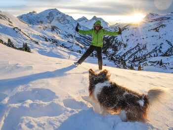 A woman with her dog on the mountain in deep snow. Behind the woman and the mountain the sun rises.