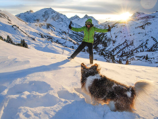 A woman with her dog on the mountain in deep snow. Behind the woman and the mountain the sun rises.
