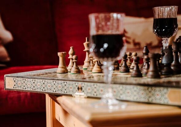 Ein kunstvoll verziertes Schachbrett steht auf einem hölzernen Tisch, daneben stehen zwei Weingläser mit Rotwein und im Hintergrund ist eine rote Samtcouch