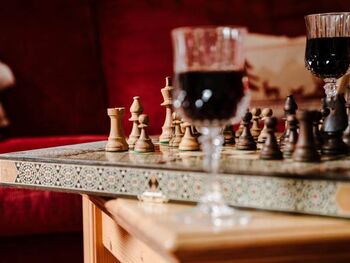 Ein kunstvoll verziertes Schachbrett steht auf einem hölzernen Tisch, daneben stehen zwei Weingläser mit Rotwein und im Hintergrund ist eine rote Samtcouch
