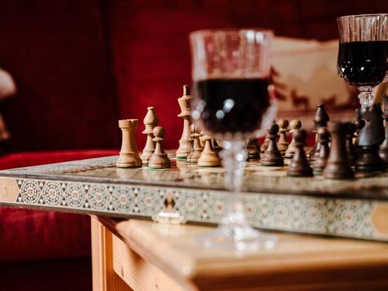 Ein kunstvoll verziertes Schachbrett steht auf einem hölzernen Tisch, daneben stehen zwei Weingläser mit Rotwein und im Hintergrund ist eine rote Samtcouch