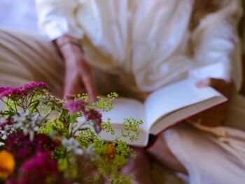 An elderly lady reading a book next to a flower bush