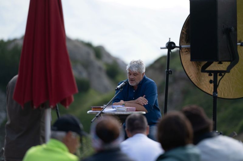 Sprecher hinter Pult vor Publikum im Hintergrund Sommer-Bergwelt